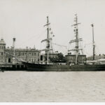 Image: A large, three-masted sailing ship is moored alongside a wharf in a port town. Several buildings, including a large nineteenth-century structure with an octagonal tower in one corner, are visible in the immediate background