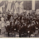 Image: Black and white photograph of a crowd of men and women, the majority of them looking at the camera