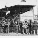 Image: Grandstand and crowds watching a horserace. The twin grandstands are full and race callers and reporters fill up the third stand