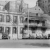 Image: 1930s era cars are parked on the street outside a two-storey stone building with shuttered windows and a balcony running halfway along the front of the building.