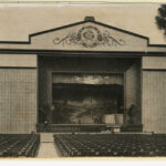 Image: a large open air theatre with rows of seats facing a stage with an outdoor scene as its backdrop