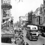 Image: 1920s and 1930s era cars, men on bicycles and a double decker bus travel down a busy city street lined with a large number of shops and a theatre