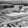 Image: An aerial photograph of a port town, at the centre of which is a large building with the words ‘General Motors Aust. Pty. Ltd’ emblazoned across it. Sailing and motor vessels of various types are visible in the river beyond the building