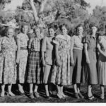 Image: A group of young Caucasian women flanked by two middle-aged Caucasian women pose for a photograph in rural central Australia. All of the women are wearing dresses of 1940s and 1950s vintage