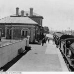 Image: A railway station. A train is on the platform to the right, with smoke coming out of the front. People are boarding the train. To the left is the station building, made of brick