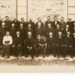 Image: Black and white group portrait. Three rows of men in suits pose against a stone building. There is one woman in the photograph. She is seated in the front row, third from left.