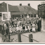 Image: A large group (over fifty) of people pose outside a hall for a group photo. The first two rows are children.