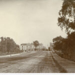 Image: a wide, muddy road stretches through parklands. In the distance the road continues through a more built-up area with numerous buildings including a corner hotel.