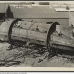 Image: The body of a tug boat upside down in the process of being constructed, numerous people, ropes and ladders surround the boat