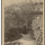 Image: Black and white photograph of a woman in front of a house