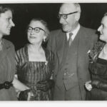 Image: A group of three middle-aged Caucasian women and one middle-aged Caucasian man speak to one another at a social event. All are wearing clothing of 1950s vintage