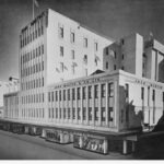 Image: a large 1930s era department store building with displays in its ground floor windows. The majority of the building is three storeys high, however, a seven storey tower protrudes from its centre.