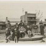 Image: A group of children sit on a merry-go-round (or joy wheel) in a park. A street and row of buildings are visible in the background.