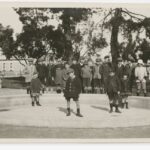 Image: A group of children pose for a photograph in an empty wading pool in a park. A street and row of buildings are visible in the background.