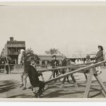 Image: A group of children play on see-saws in a park. A street and row of buildings are visible in the background