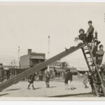 Image: A small group of children play on a slide in a park. A street and row of buildings are visible in the background