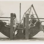 Image: Two boys sit in a ‘rocking boat’ at a playground. A swings-set is positioned immediately behind the boys