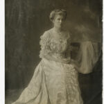 Image: Photographic portrait of a young Caucasian woman in Edwardian-era attire. She is seated and wearing a floor-length light-coloured dress with embroidered decoration