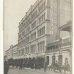 Image: a large number of people in early 20th century clothing gather outside a five storey building with a decorative iron verandah