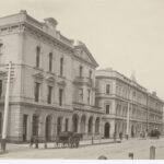 Image: A row of three storey grand stone buildings lining a dirt street.