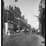 Image: A wide dirt road with a number of pedestrians and horse drawn vehicles. The buildings are decorated with flags and greenery. One of the buildings on the left is still under construction and is covered in scaffolding.