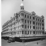 Image: a large five storey building with a verandah situated on a street corner. The corner of the building is rounded and extends into a cupola with a flagpole. A double decker bus passes on the street outside while a policeman stands directing traffic.