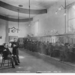 Image: Interior view of a telephone exchange. A row of women switchboard operators are supervised by a well dressed woman sitting at a raised desk on the left.