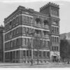 Image: a four storey brick building with an adjoining five storey crenelated tower in one corner.  There are recessed balconies on the third and fourth floors.