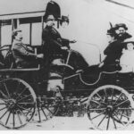 Image: A small group of Caucasian men, women and children dressed in late Victorian attire ride in a carriage-like vehicle along a town road. The vehicle has four spoked wheels and what appears to be an engine. A moustachioed middle-aged man operates it