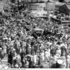 Image: Four 1940s-era auto-mobiles move through a crowd of people gathered for a ceremony