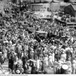 Image: Four 1940s-era auto-mobiles move through a crowd of people gathered for a ceremony