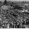 Image: A large group of people dressed in 1940s attire gather on the approach to a bridge. Several buildings are visible just beyond the bridge approach