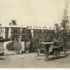 Image: a horse drawn carriage is pulled up alongside large gates. Beyond the gates a long drive curves around to meet a large two storey stone building with arched windows and a large number of chimneys.