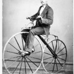 Image: man in a top hat and suit riding a velocipede with wooden wheels