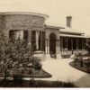 Image: a small number of women and girls in 1860s era clothing stand outside a single storey bluestone house with curved wall, columned portico and verandah.