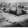 Image: a high angle view of a low lying city. In the foreground, situated at a crossroad, is a stone church with a single square tower with crenellations, which becomes octagonal with a very steeply pitched roof, to one side  of the building