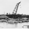 Image: A large derrick crane sits adjacent to one approach to a concrete bridge span. The other half of the bridge, also under construction, is visible in the background