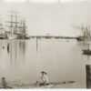 Image: A young Caucasian boy in late nineteenth century attire sits on a spar with his legs dangling in a river. Several sailing ships, small steamships, and a bridge are visible in the background