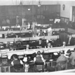 Image: The inside of a telegraph office. Men sit at rows of desks with telegraph machines