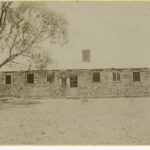 Image: Front view of a long stone building with barred windows and a tree in front.