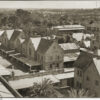 Image: an aerial view of a complex of one and two storey stone buildings with slate and tin roofs including one with verandahs under which 1930s cars are parked.