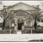 Image: Black and white photograph of the front of a building. Palm trees stand on either side of the front of the building. The building reads "ART GALLERY"