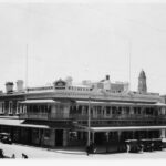 Image: A bluestone rubble hotel, with a chamfered corner entrance and balcony extending across its front face and around one side. A number of 1920s cars are parked on the street outside.