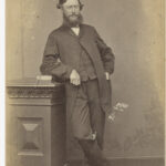 Image: bearded man in suit leaning against pedestal with book on it