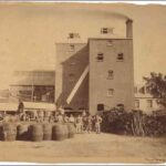 Image: a group of men in late 19th century worker's clothes stand with a collection of large barrels outside of a tall red brick brewery building.