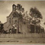 Image: A large stone church bordered by a wooden picket fence and a handful of trees. Part of a dirt street is visible in the foreground