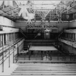 Image: A large, empty swimming pool in a vast open building with a roof. A wrestling ring is positioned within the empty pool