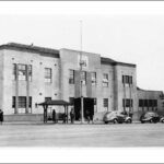 Image: A two-storey building with plain façade at the intersection of two streets. 1940s-era auto-mobiles and pedestrians are on the street in front of the building