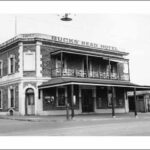 Image: a two storey corner hotel with a small balcony on one side. A verandah protrudes further out to the street beneath the balcony. A parapet sign reads: "TISHERS BUCKS' HEAD HOTEL"