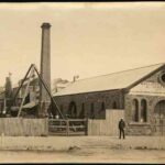 Image: a man in 1870s era clothing stands outside a small stone foundry with arched windows and doors and a medium pitched gable roof. To the left of the building is a fenced yard where more men work at various machinery next to a tall brick chimney.